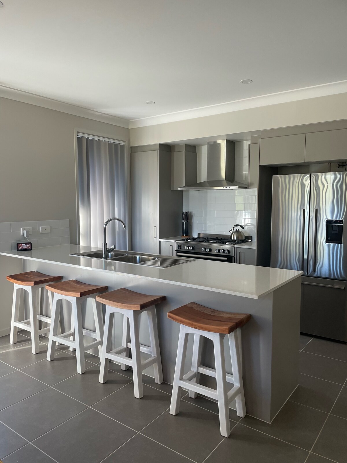 The open-plan kitchen features modern appliances, including a double oven and five hobs. A spacious island with sleek countertops is complemented by four wooden stools, creating an inviting space for meal preparation and gatherings. Natural light filters through the nearby windows, enhancing the airy atmosphere.