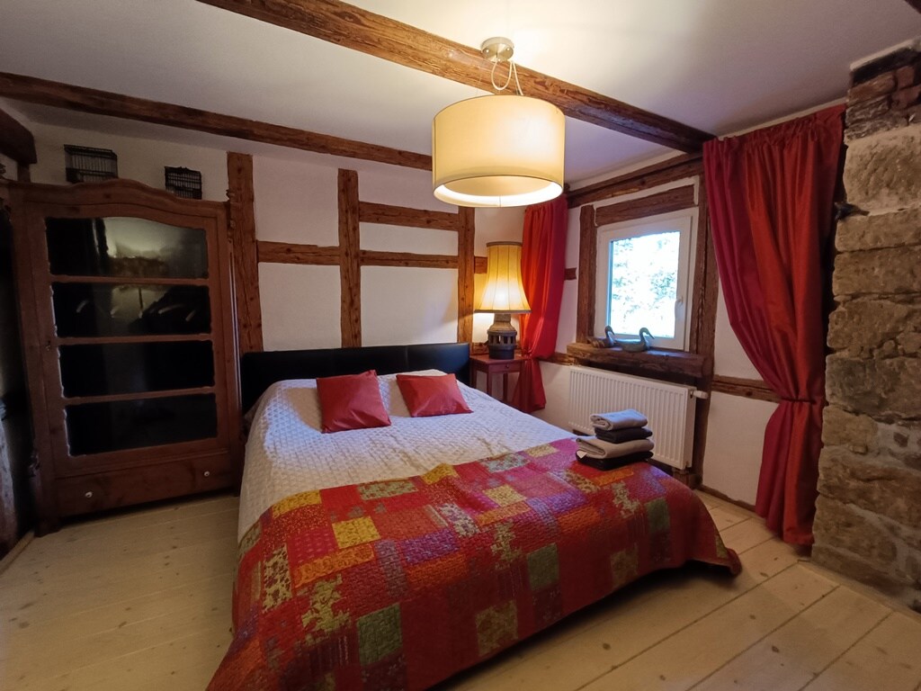 A well-appointed room features a spacious bed with a colorful quilt and two decorative pillows. Natural light enters through a window, complemented by warm red curtains. An elegant lamp provides additional lighting, and a wooden cabinet enhances the rustic charm of the decor.