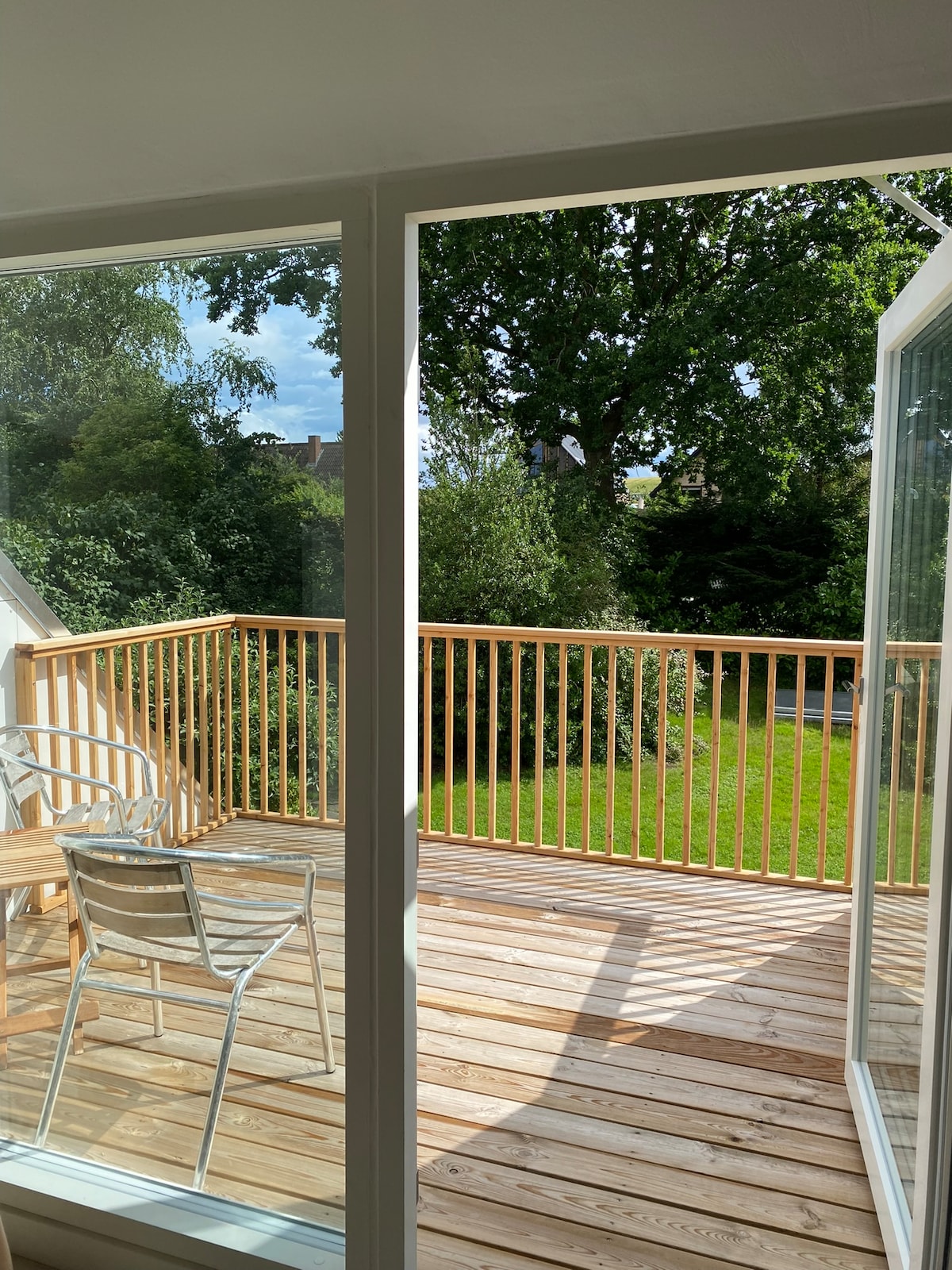 A view of the private terrace features a wooden railing and natural wood flooring. Two metal chairs are positioned for seating. Lush greenery and a well-maintained lawn are visible beyond the terrace, enhancing the sense of outdoor space.