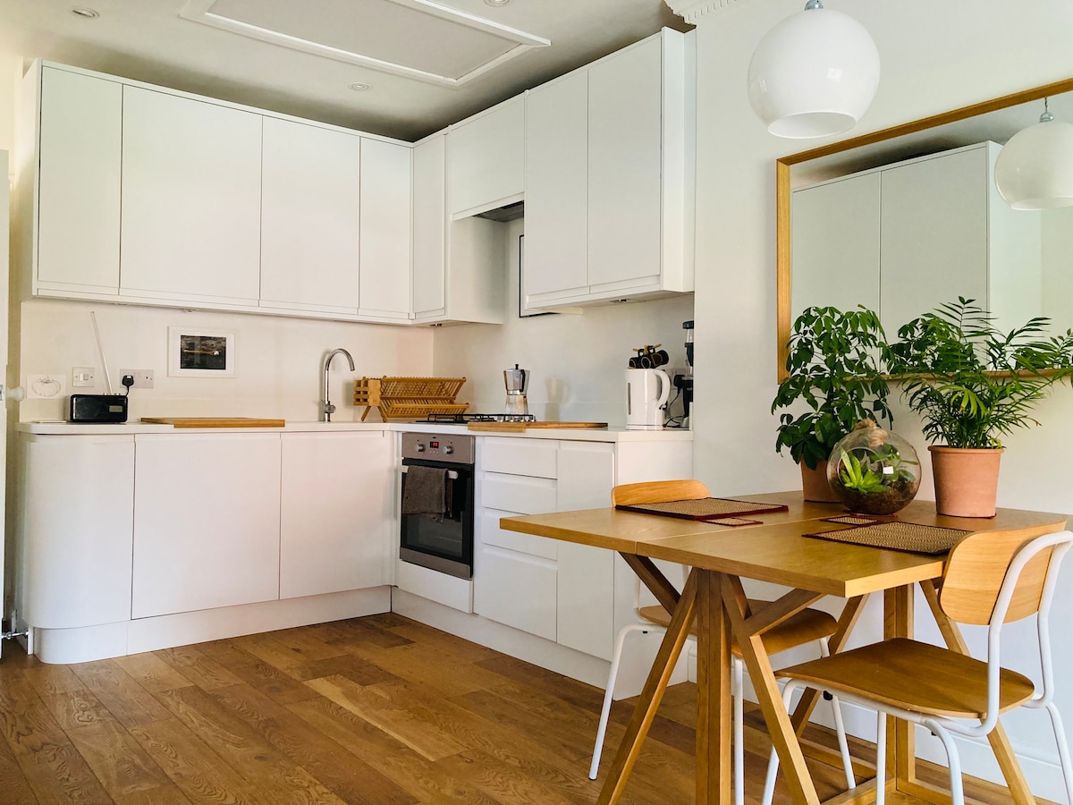 A modern kitchen area is displayed with white cabinetry and a wooden countertop. A dining table with four wooden chairs sits next to a small indoor plant. Essentials such as a kettle and coffee machine are visible, enhancing the functional space.