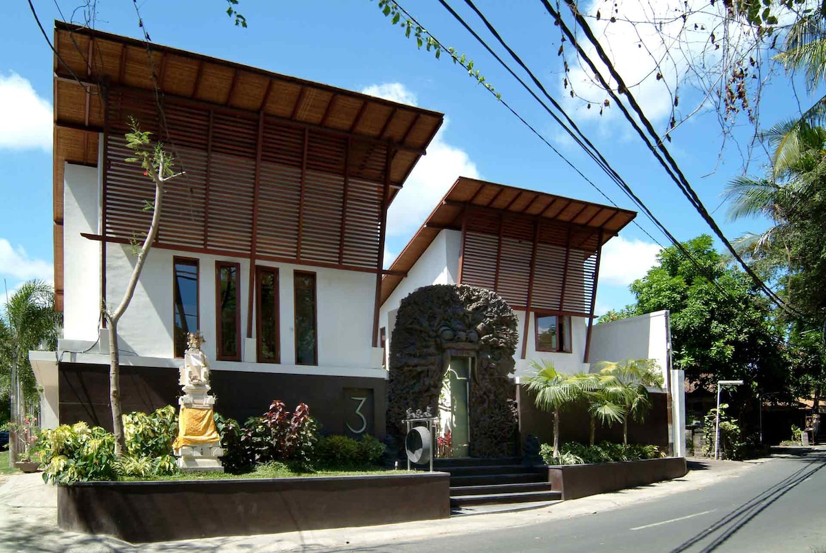 The villa's exterior showcases a modern architectural design with large windows and a wooden roof. Lush greenery complements the property, with decorative stonework framing the entrance. Bright sunlight illuminates the facade, creating an inviting atmosphere in the tropical setting.