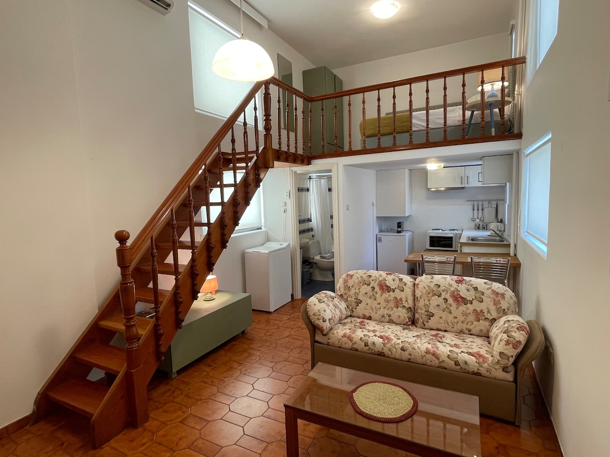 The open-plan living area features a floral-patterned sofa and a wooden coffee table. A staircase leads to a lofted sleeping area, while the kitchen with modern appliances is visible in the background. Natural light filters through windows, highlighting the warm tones of the tiled floor.