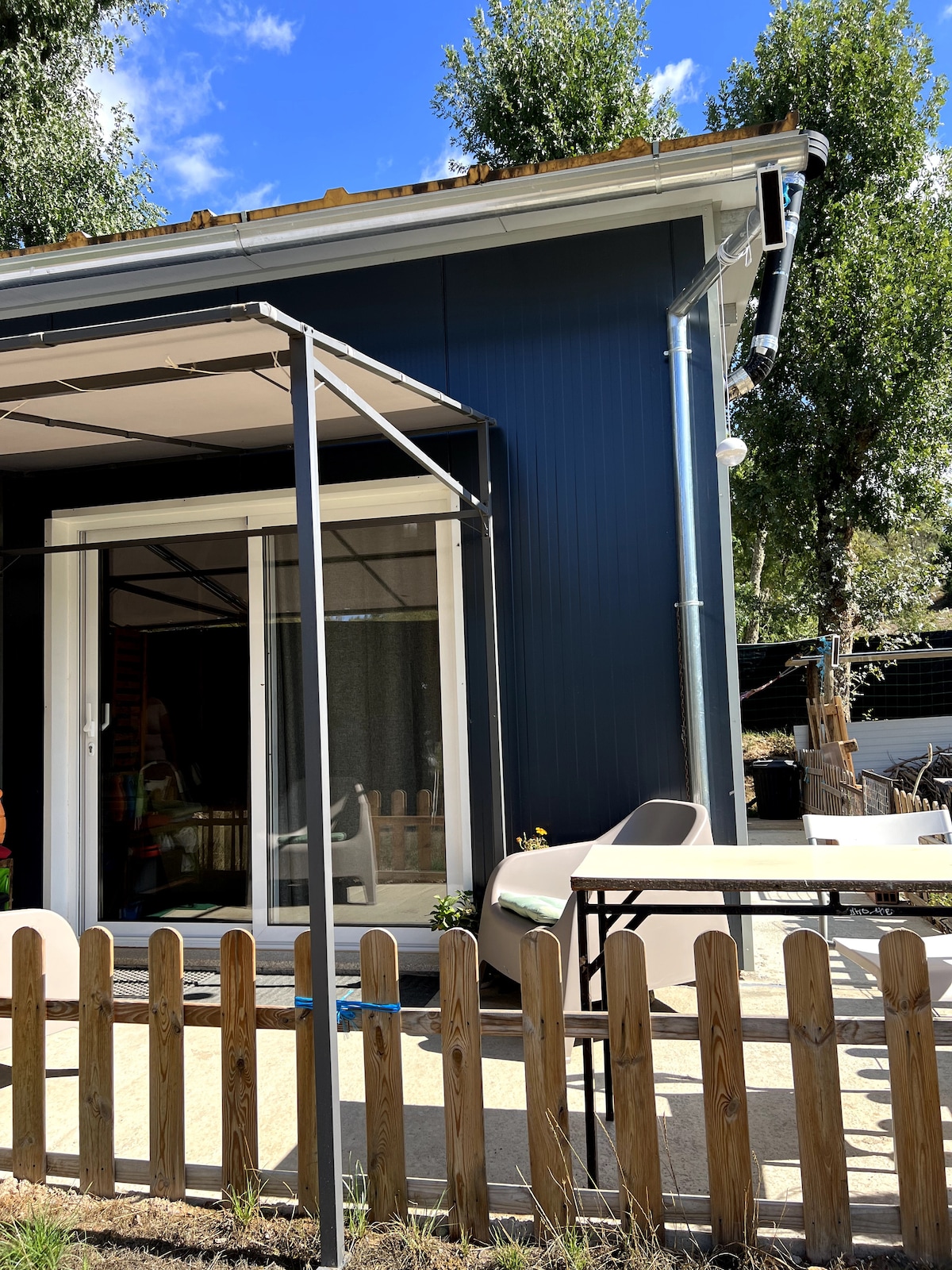 A modern mini house is presented with a blue exterior, featuring large glass doors that open to a small patio area. A simple table and chairs are positioned on the patio, surrounded by a wooden fence that adds to the outdoor ambiance. Bright trees are visible in the background.