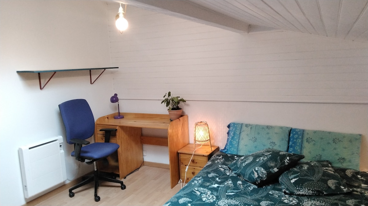 A well-lit bedroom features a spacious bed adorned with patterned bedding. A work desk and chair are positioned beside a small plant, while a decorative lamp adds warmth. The room's walls are painted white, and the ceiling showcases wooden beams.