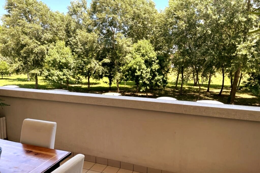 A spacious terrace features a wooden table surrounded by white chairs. A view of lush green trees creates a serene backdrop, enhancing the calm atmosphere. The flooring is tiled, and the terrace walls are low, offering an unobstructed connection to the outdoors.