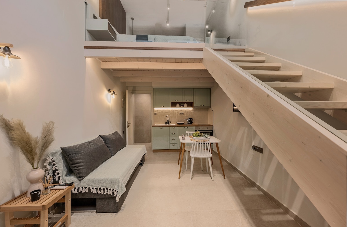 An interior space features a light wood staircase leading to an upper level. A cozy sofa with a throw is positioned near a small dining table. The kitchenette showcases modern appliances and an inviting color palette. Soft lighting enhances the warm ambiance.