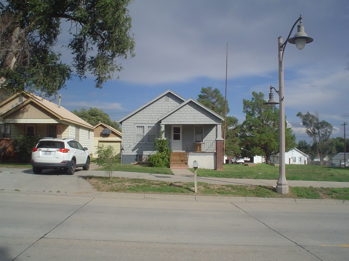A single-story house with a gray shingle exterior is positioned in a suburban setting. The front features a small porch and a yard with grass. A white vehicle is parked along the side of the house, while a street light and neighboring homes are visible in the background.