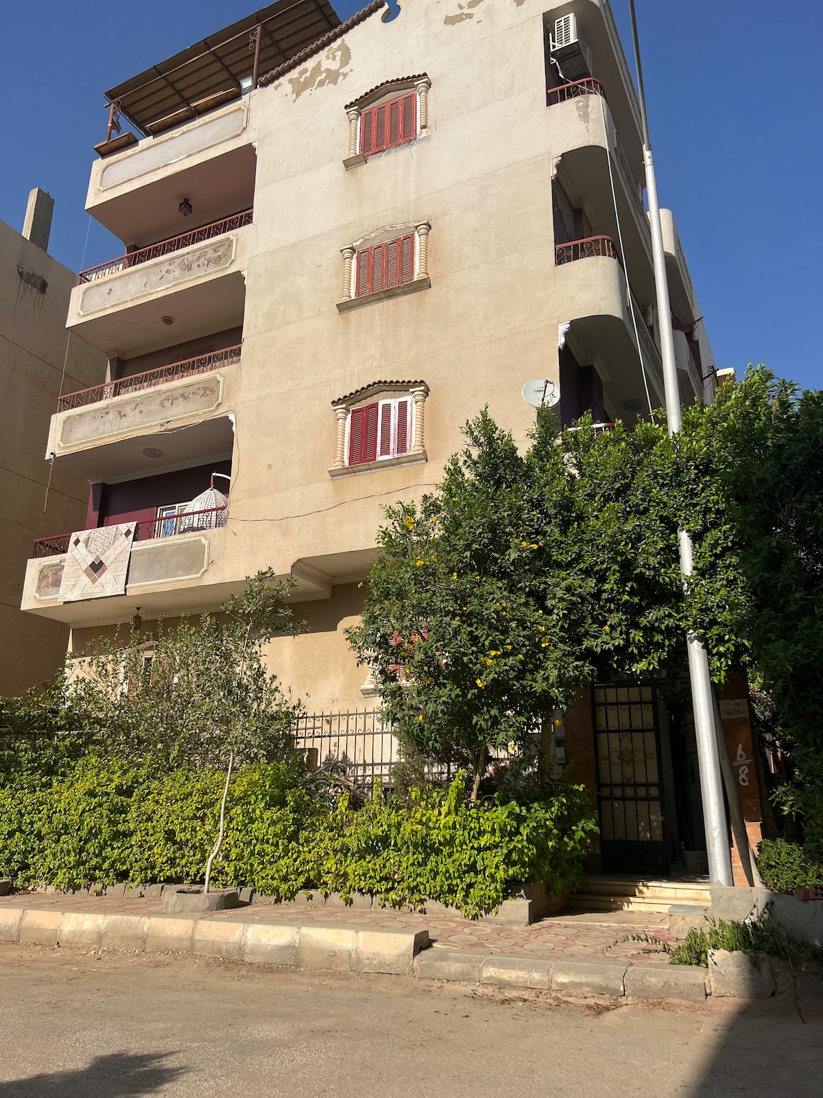 A three-story building features a beige façade with balconies and red-tinted windows. Lush greenery lines the entrance, with a pathway leading to the gated front door. The building is set against a clear blue sky, indicating a sunny day.
