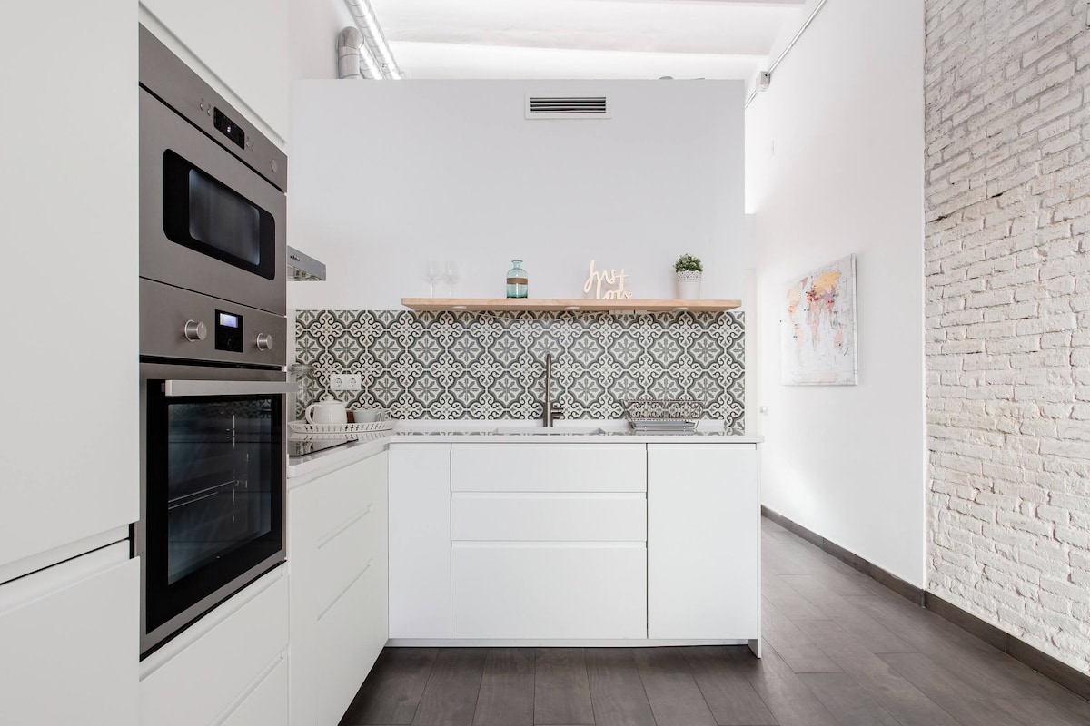 A modern kitchen is visible, featuring sleek white cabinetry and stylish tile backsplash with intricate patterns. Stainless steel appliances, including an oven and microwave, integrate seamlessly into the design. Natural light enhances the space, highlighting the minimalist aesthetic.