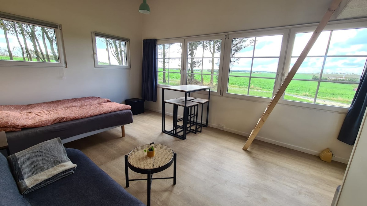 A spacious interior is highlighted by large windows that offer views of the surrounding fields. A three-quarter bed and a sofa are positioned within the room. A small table paired with two chairs is arranged near the window, enhancing the natural light in the space.