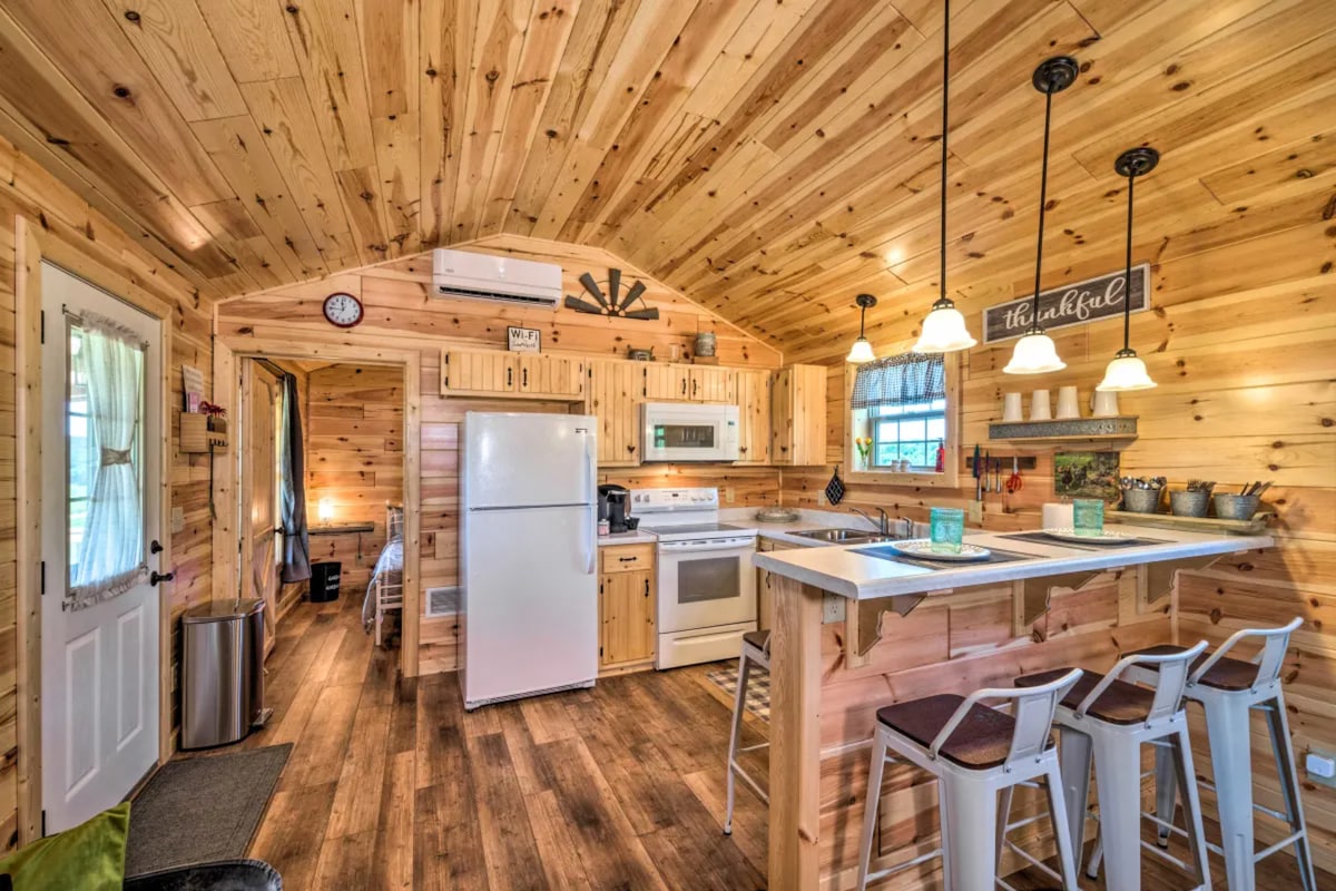 The kitchen area features natural wood paneling and an open layout, equipped with modern appliances including a refrigerator, oven, and microwave. A countertop with seating for three is visible, alongside light fixtures that provide warm illumination. A door leads to the outdoors, enhancing the inviting atmosphere.