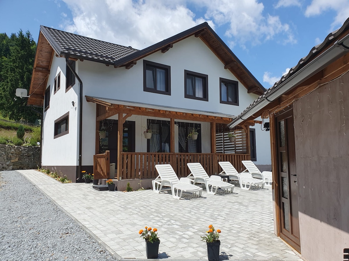 The exterior of a spacious villa is shown, featuring a covered porch adorned with hanging flower baskets. Sun loungers are arranged on a stone patio, surrounded by a well-maintained gravel area. A landscaped yard with flower pots enhances the inviting entrance.