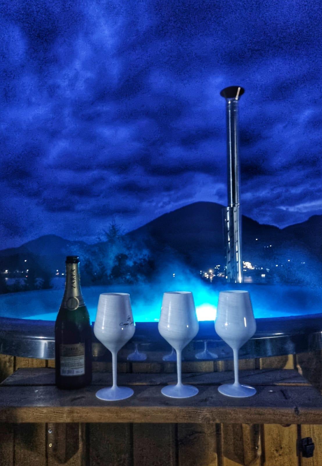 A bubbling hot tub is set against a dramatic night sky, complemented by soft blue lights. Three elegant glasses stand ready on a wooden surface beside a bottle of sparkling wine, creating a serene ambiance for relaxation.