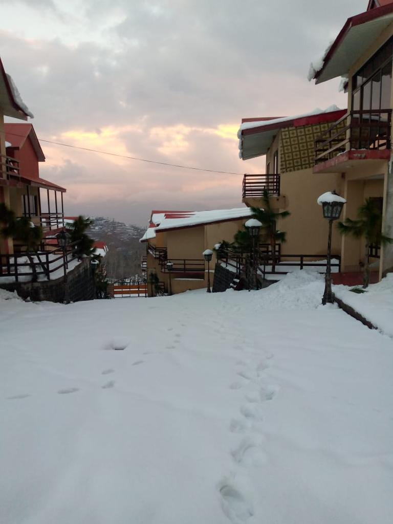 A snow-covered pathway winds through a charming arrangement of chalet-style buildings, framed by trees and decorative lamp posts. The sky above features soft pastel colors, hinting at sunrise or sunset, while the distant mountains offer a serene backdrop to this peaceful setting.
