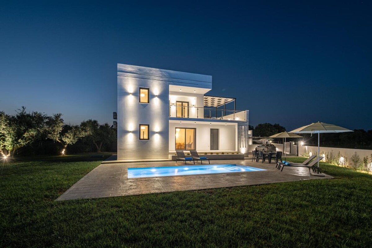 The modern villa is illuminated at night, showcasing its sleek white exterior and expansive layout. The private swimming pool glows under the soft lighting, surrounded by a manicured lawn. Outdoor furniture is positioned adjacent to the pool, with trees lining the background adding to the serene ambiance.