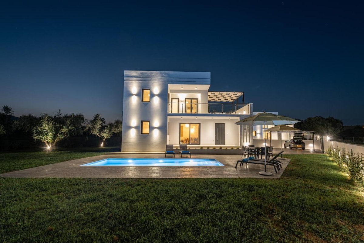 A modern villa is depicted at night, illuminated by soft exterior lighting. The private swimming pool reflects the surrounding greenery. Outdoor furniture, including loungers and umbrellas, is arranged near the pool. A spacious terrace offers access to the elegantly designed interior.