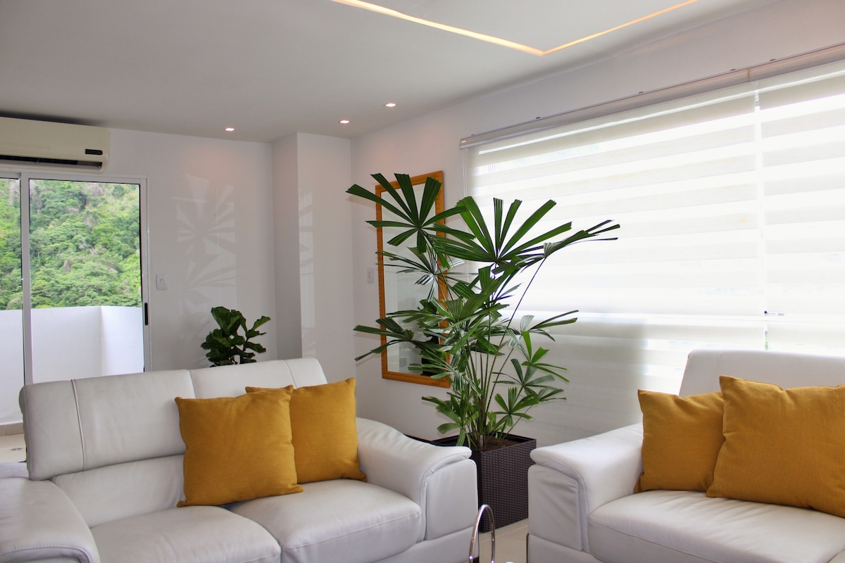 A bright and airy living space features white sofas with yellow cushions, alongside lush green plants. Natural light filters through sheer window coverings, illuminating the room. A large wall mirror reflects the vibe, enhancing the sense of openness and tranquility.