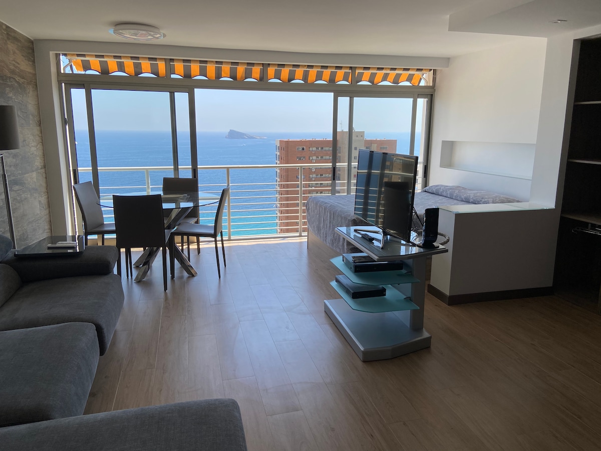 A modern loft interior is featured, with a spacious living area that includes a comfortable seating arrangement and a sleek television stand. Floor-to-ceiling windows provide unobstructed views of the ocean, while a dining table for four is situated near the glass doors.