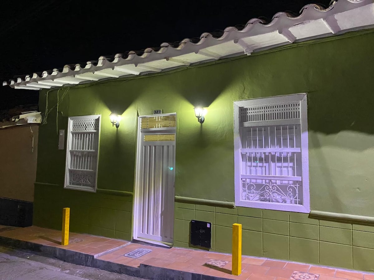 The exterior of the house is illuminated at night, showcasing a vibrant green facade. Two wall-mounted lights flank the front entrance, highlighting the traditional wooden windows with intricate designs. The tiled pathway is bordered by yellow posts, enhancing the welcoming appearance of the home.