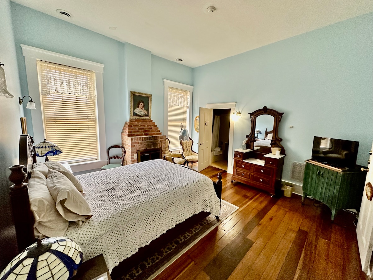 A spacious room features a queen bed with a textured white coverlet, complemented by traditional wooden furniture. Natural light enters through two windows with soft shades. A vintage fireplace adds charm, while a flatscreen TV sits on a wooden dresser, and a private bathroom is visible.