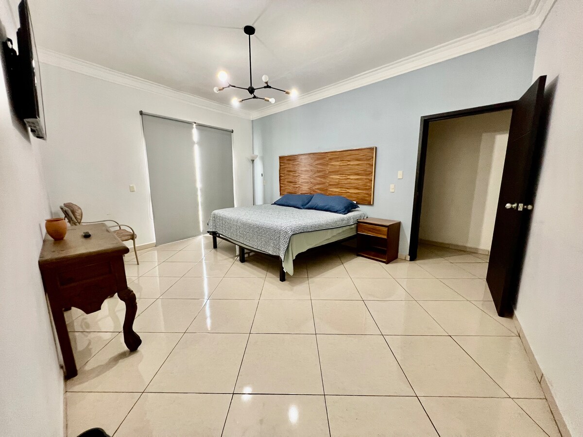 A private room is shown, featuring a spacious bed with a blue coverlet. Light enters through a window, illuminating the tiled floor. A wooden nightstand and an antique-style desk are positioned nearby, complementing the minimalistic design.