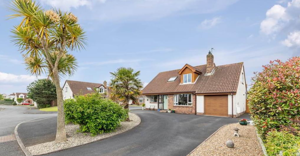 The exterior of a large family vacation home is shown, featuring a well-maintained driveway leading to the front entrance. Lush greenery and palm trees are present, adding a touch of tropical ambiance. The home is set in a quiet neighborhood with ample space around it.