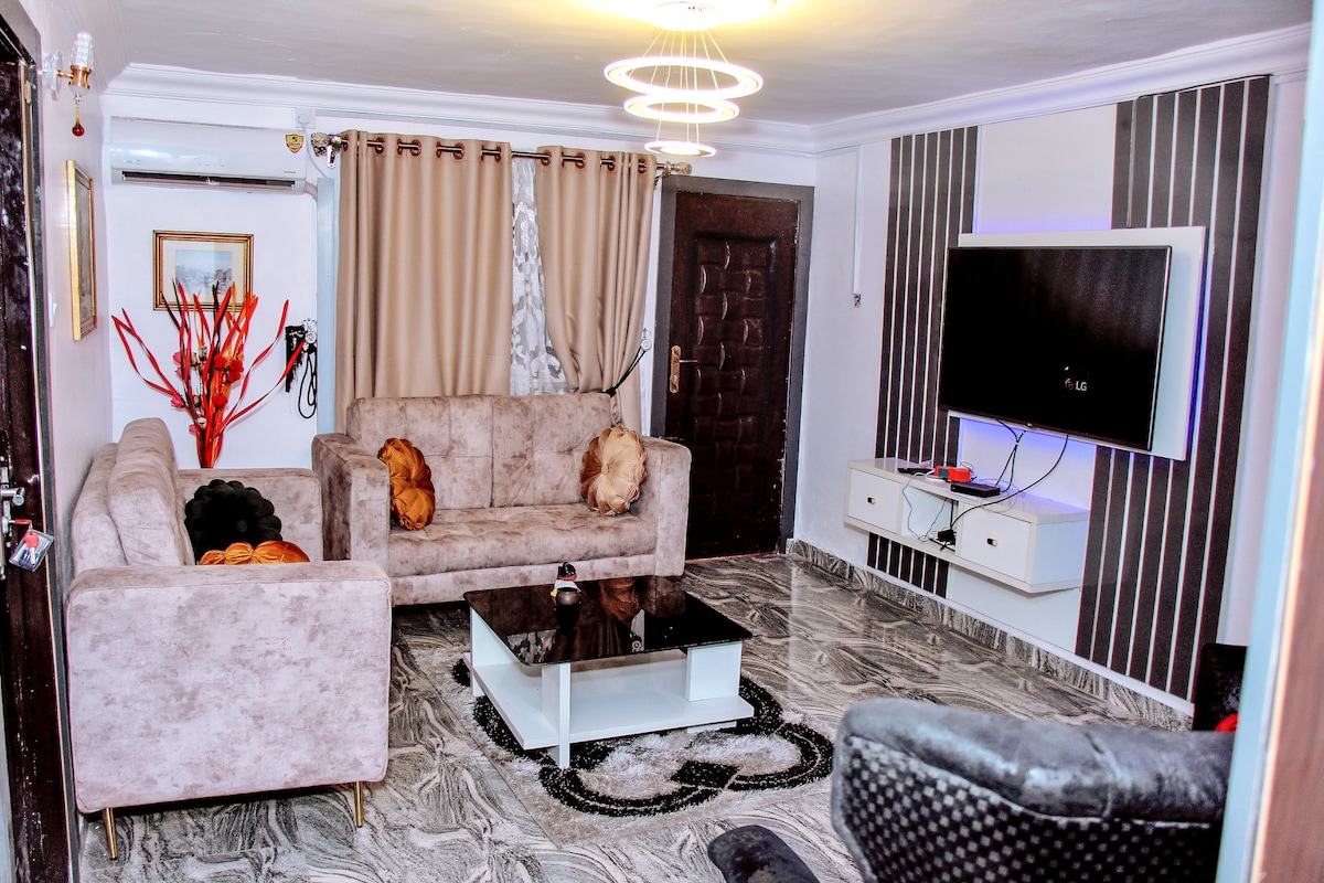 A spacious living room features two light-colored sofas arranged around a black and white coffee table. A large television is mounted on a stylish feature wall, complemented by elegant curtains. A decorative plant adds a touch of color, while air conditioning is also visible.