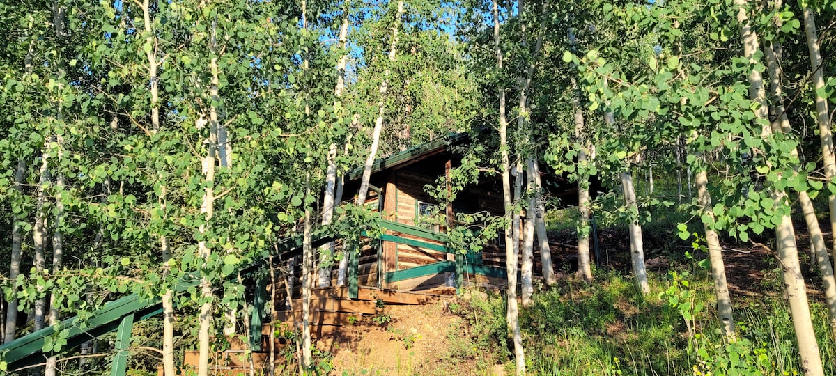 A rustic log cabin is nestled among lush green aspen trees, creating a private and serene atmosphere. Sunlight filters through the leaves, illuminating the cabin's wooden exterior, which blends harmoniously with the surrounding nature.