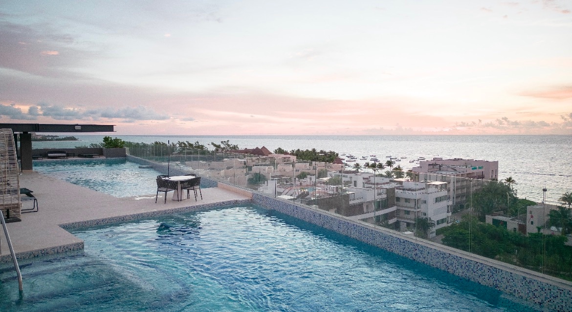 A rooftop pool area is presented, featuring two tranquil swimming pools surrounded by a serene landscape. Comfortable seating is positioned beside the water, with panoramic views of the ocean and nearby buildings. The sky displays soft pastel hues of sunset, enhancing the calming environment.