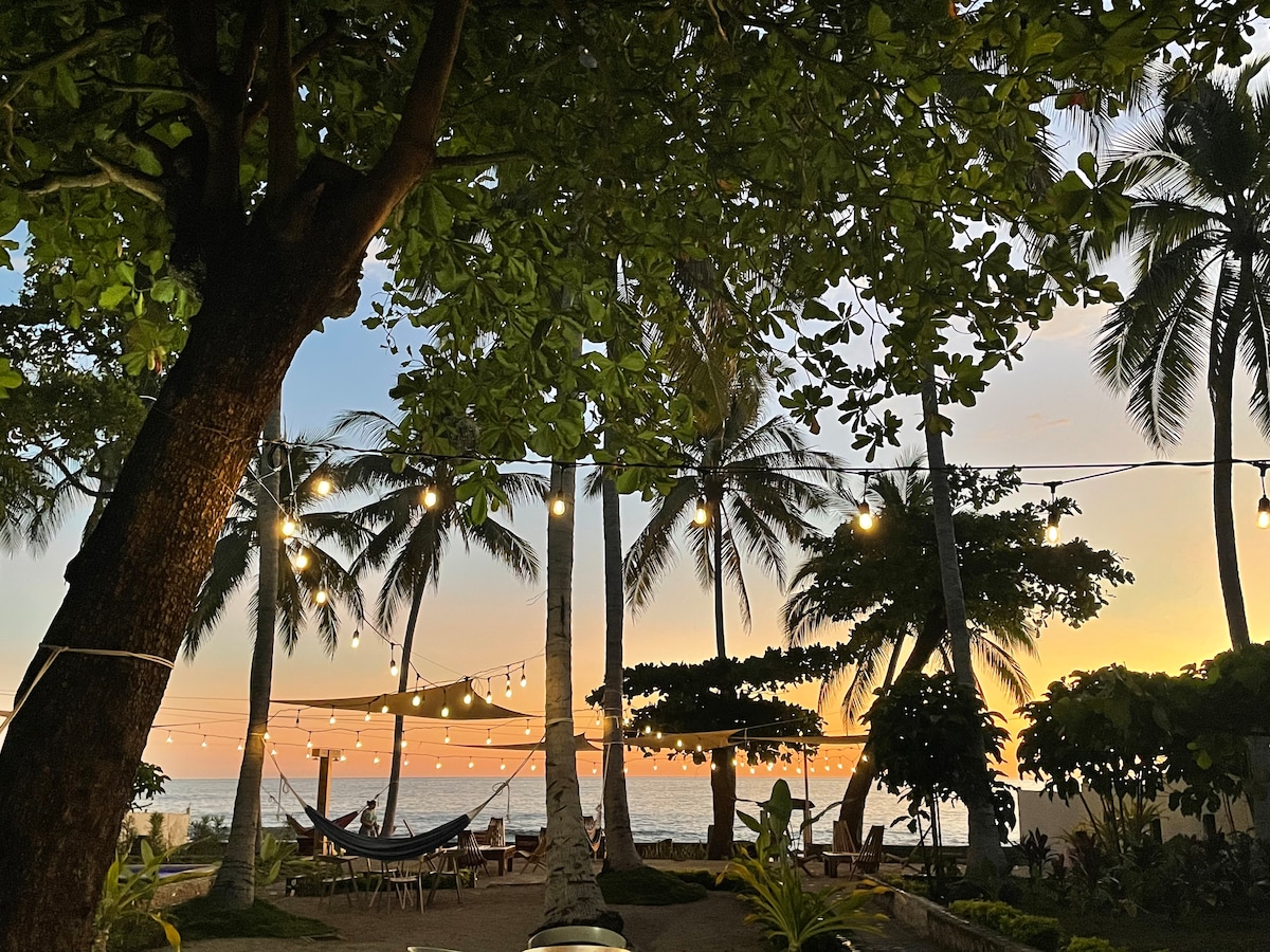 A serene outdoor space is framed by tall palm trees and illuminated by soft string lights. A hammock is positioned between two trees, with a view of the ocean in the background. The warm glow of the sunset casts an inviting light across the area.