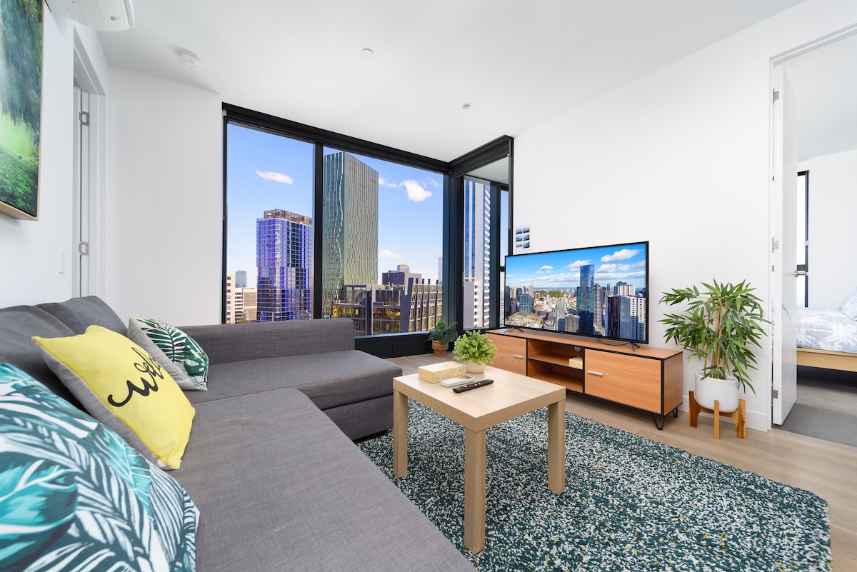 A modern living area features a comfortable gray sofa and a stylish coffee table. A large television displays a vibrant city skyline through expansive windows, with natural light illuminating the space. Greenery adds a touch of nature, enhancing the contemporary design.