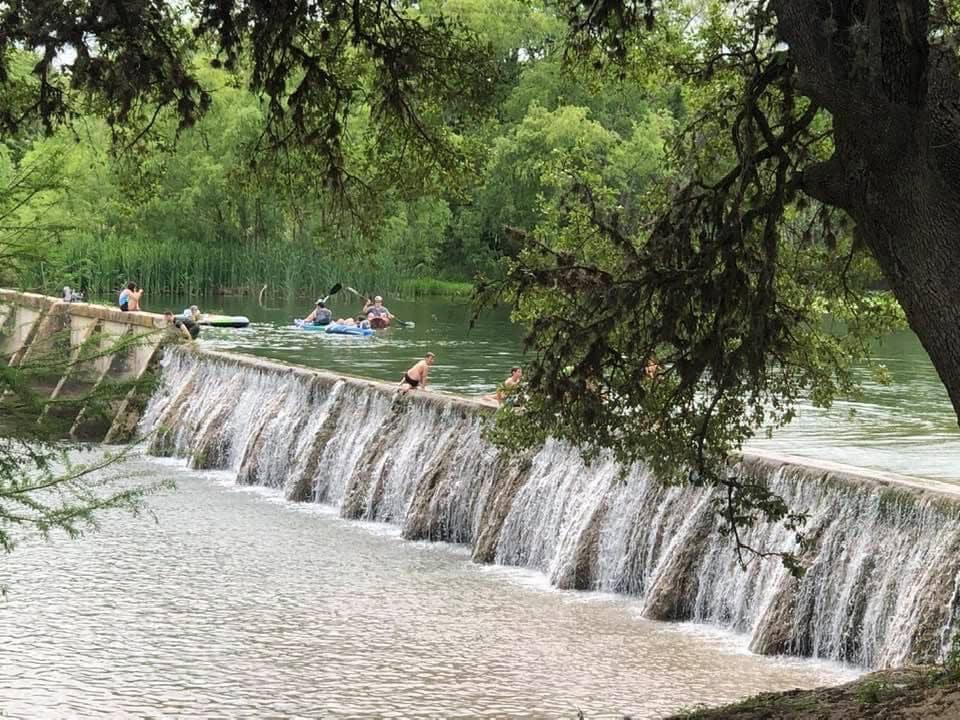 A serene view showcases a cascading waterfall flowing into a tranquil river. Individuals can be seen enjoying recreational activities in the water, surrounded by lush green foliage and distant reeds. The scene reflects a harmonious blend of nature and leisure.