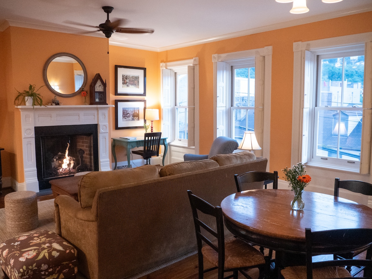 An inviting living area features a cozy brown sofa facing a gas log fireplace, surrounded by windows that allow natural light. A wooden dining table invites gatherings, complemented by a decorative floral arrangement. Warm orange walls enhance the home's welcoming atmosphere.