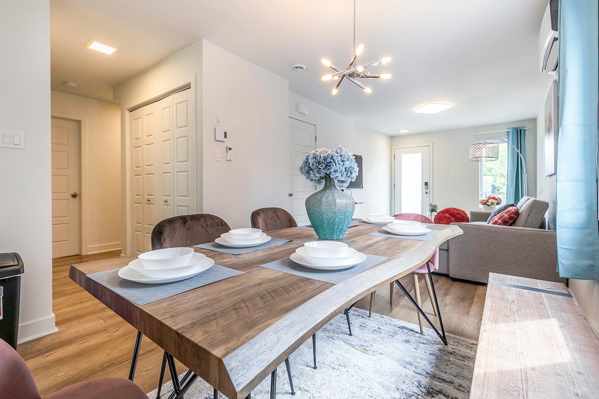 A modern dining area features a wood dining table set with plates and a decorative vase filled with flowers. A spacious living area is visible in the background, showcasing a cozy seating arrangement and natural light coming from a nearby door.
