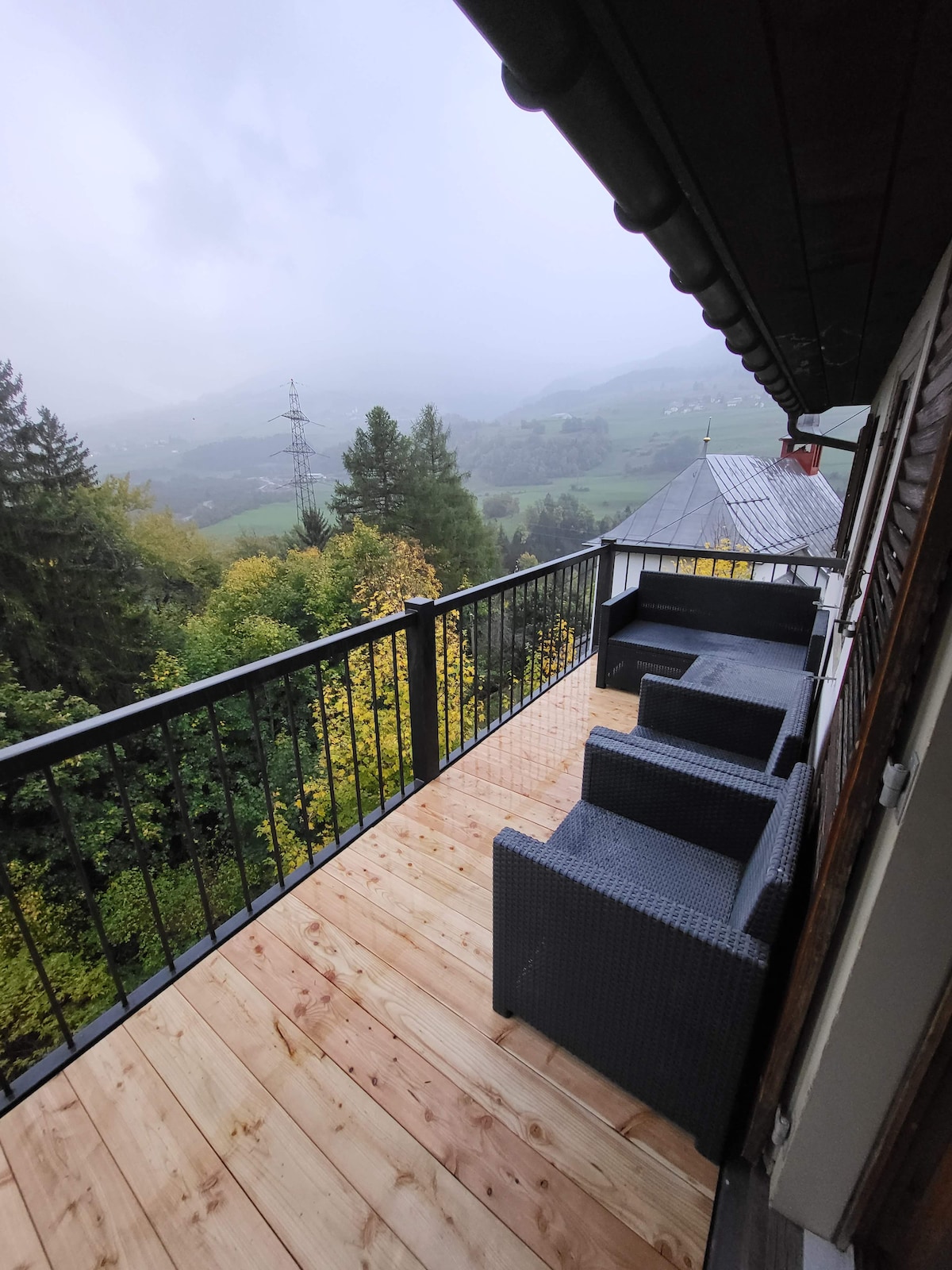 A spacious balcony is shown, featuring two comfortable chairs made of dark wicker. The wooden deck provides an inviting space to relax, while a serene landscape of misty hills and trees unfolds in the background.