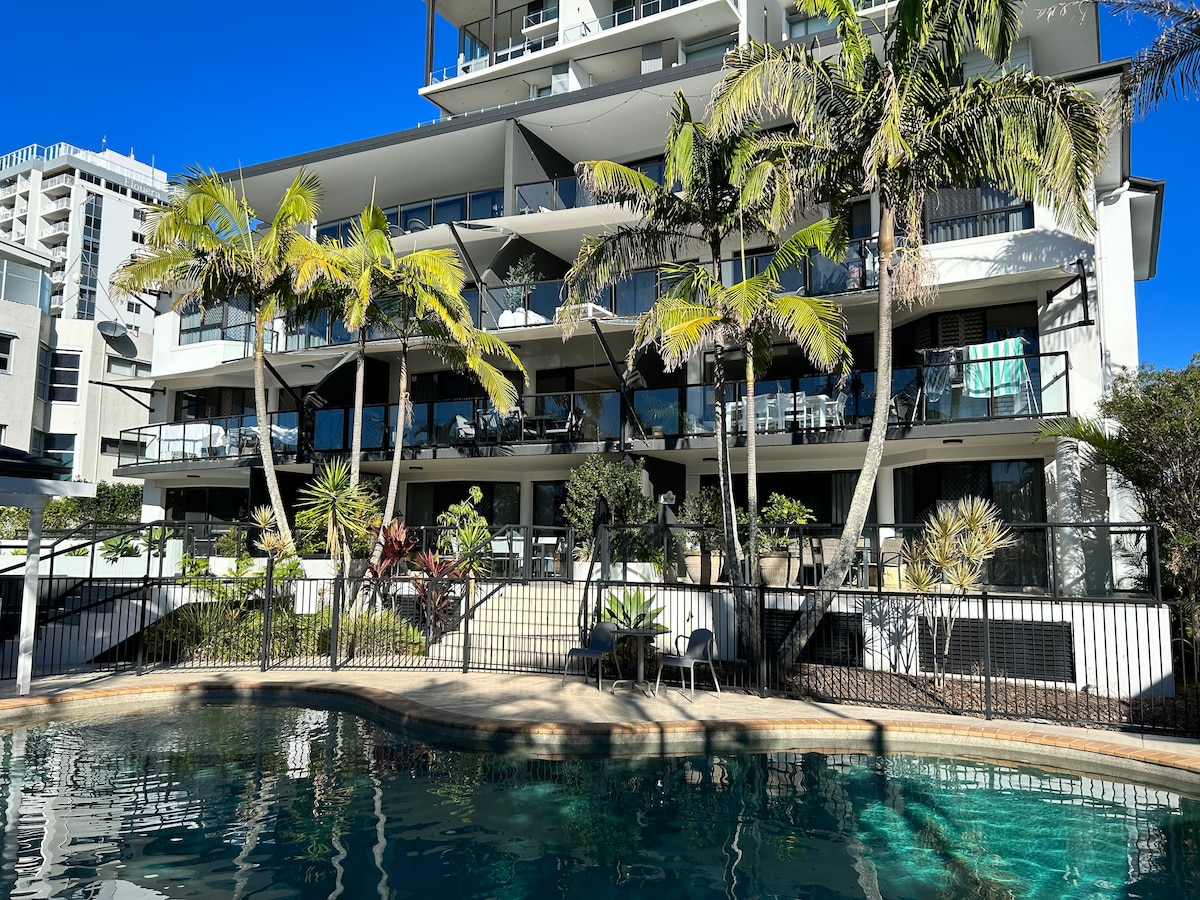 The modern low-rise complex features multiple balconies surrounded by palm trees, overlooking a clear swimming pool. The building’s architecture showcases a blend of glass and concrete, with shaded outdoor areas. Sunlight illuminates the space, creating a vibrant and inviting environment.