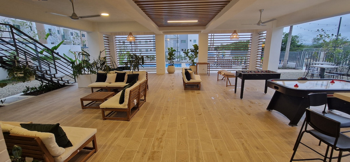 An open common area features wooden flooring and a ceiling with wooden beams. Comfortable seating is arranged with plush cushions, while a game table and foosball table are positioned nearby. Large windows and greenery enhance the space, providing a relaxed atmosphere.