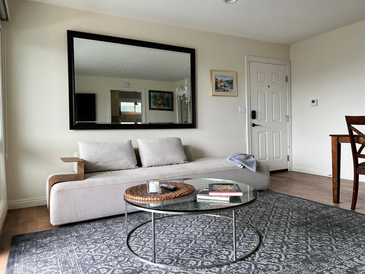 A spacious living area features a light-colored, modern sofa positioned near a large mirror. A round glass coffee table rests on a patterned area rug, while a wooden dining table is visible in the background. Natural light fills the space, highlighting decorative wall art.