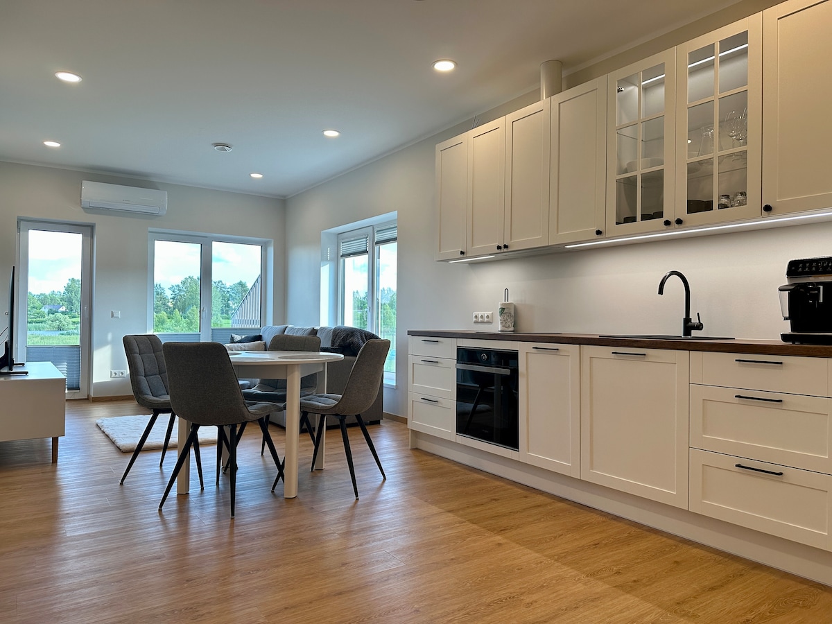 An open kitchen-living area is presented with modern cabinetry and a dining table surrounded by four grey chairs. A large window offers views of the outside greenery, while a television is visible in the corner. Soft lighting enhances the spacious feel of the room.
