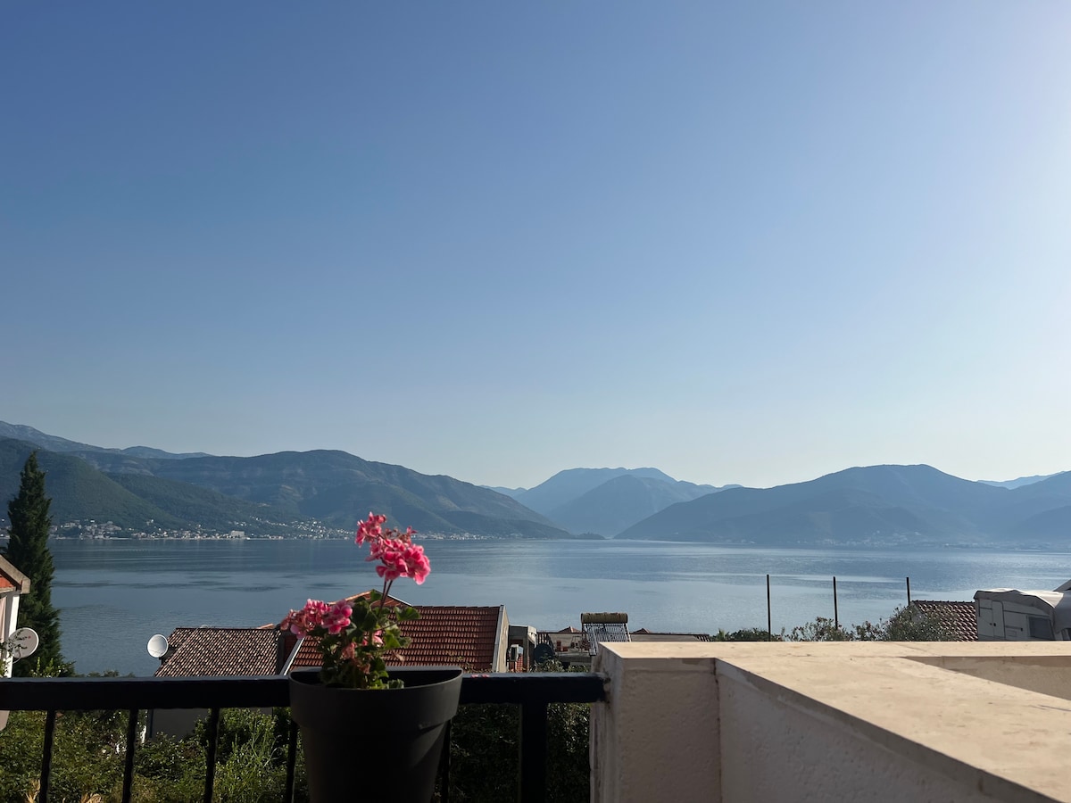 A serene view of Boka Bay is visible from the terrace, with gentle mountains encircling the horizon. A potted pink flower adds a touch of color in the foreground, while the calm water reflects the clear sky.