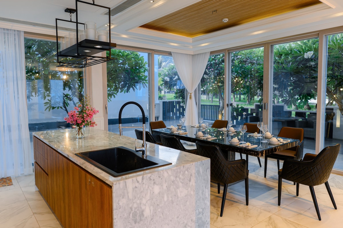 A modern dining area showcases an elegant table set for meals, surrounded by stylish chairs. Natural light filters through large windows, illuminating the space. A sleek kitchen island with a black sink complements the design, while floral decor adds a touch of freshness.