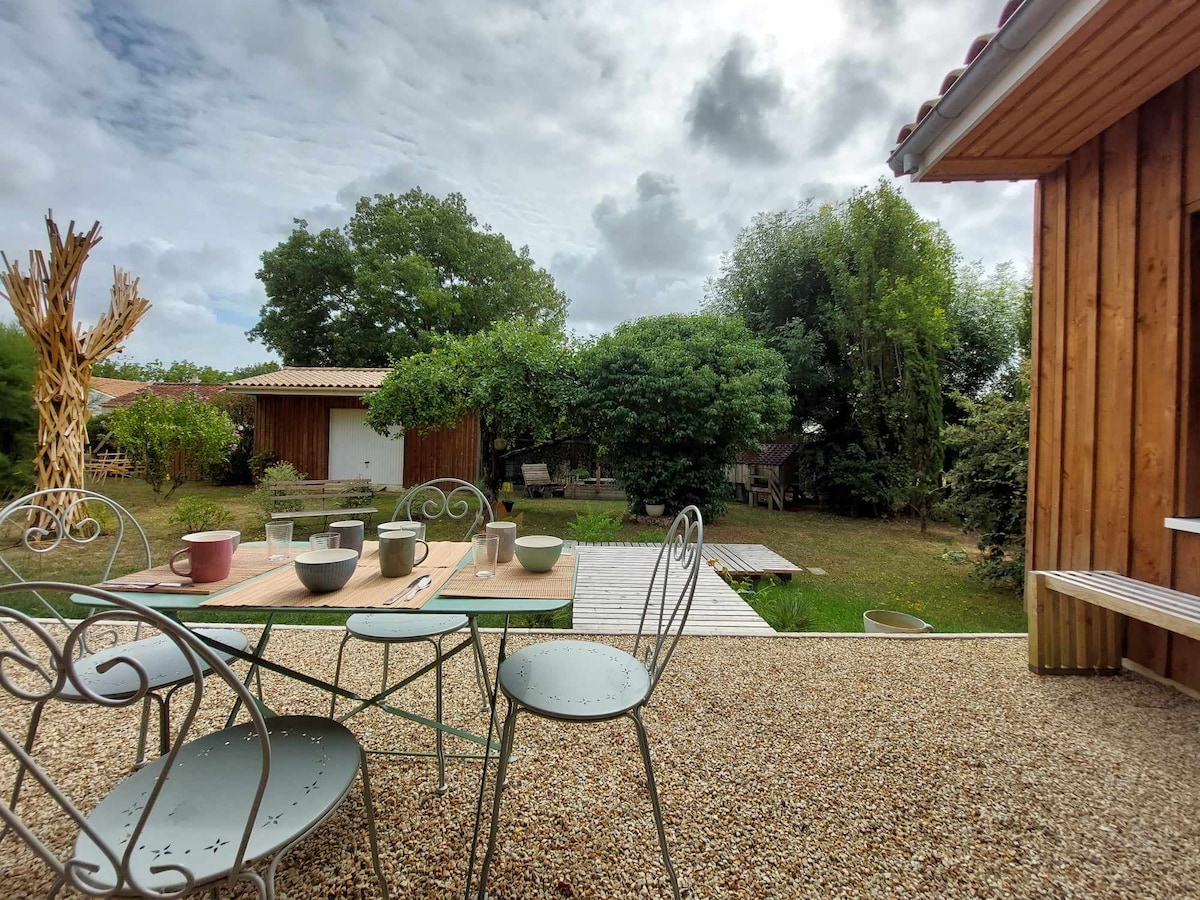 A dining area is set outdoors with a light-colored table and metal chairs, surrounded by a gravel patio. A variety of plates and cups are arranged on the table. In the background, a lush garden with trees and a wooden structure is visible.