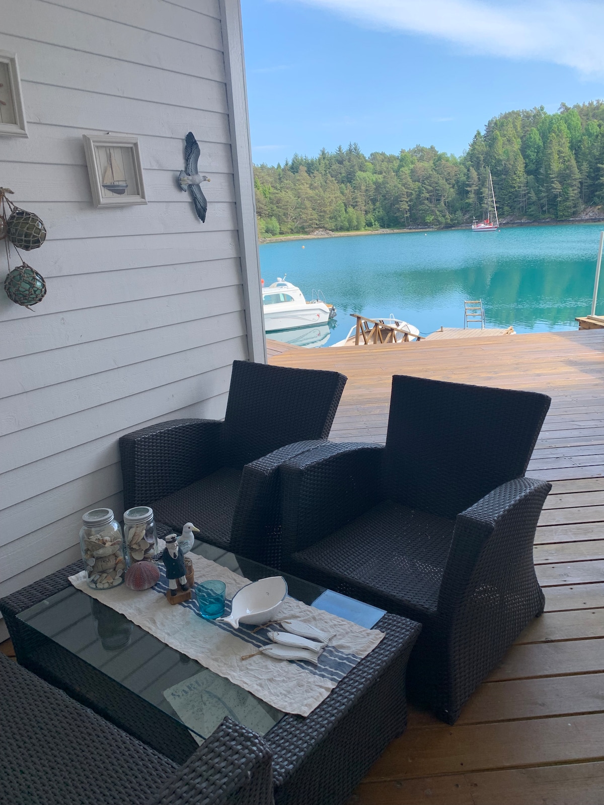 A cozy outdoor seating area is featured on a wooden deck, overlooking a tranquil body of water. Two wicker chairs are placed around a glass-topped table, decorated with seashells and coastal décor. Lush greenery surrounds the serene setting.