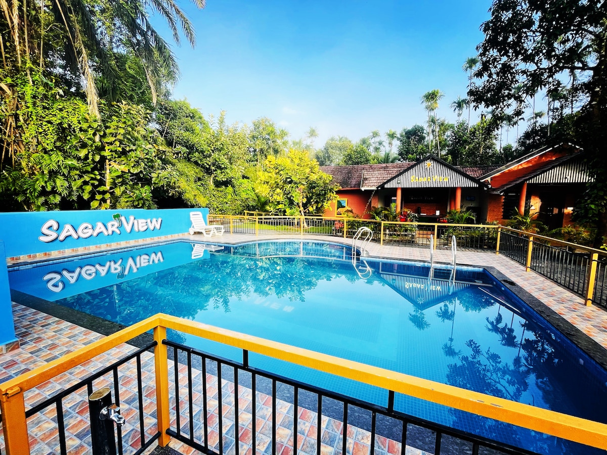 A clear swimming pool is framed by vibrant greenery and colorful tiles. A poolside area features a blue lounge chair and sturdy railings for safety. The resort's building, visible in the background, blends harmoniously with the natural surroundings.