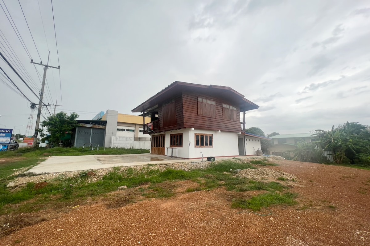 A two-story wooden house is showcased in a rural setting, surrounded by greenery and gravel. Large windows and a balcony provide natural light, while an ample parking space is present in front. The sky is overcast, adding a calm atmosphere to the scene.