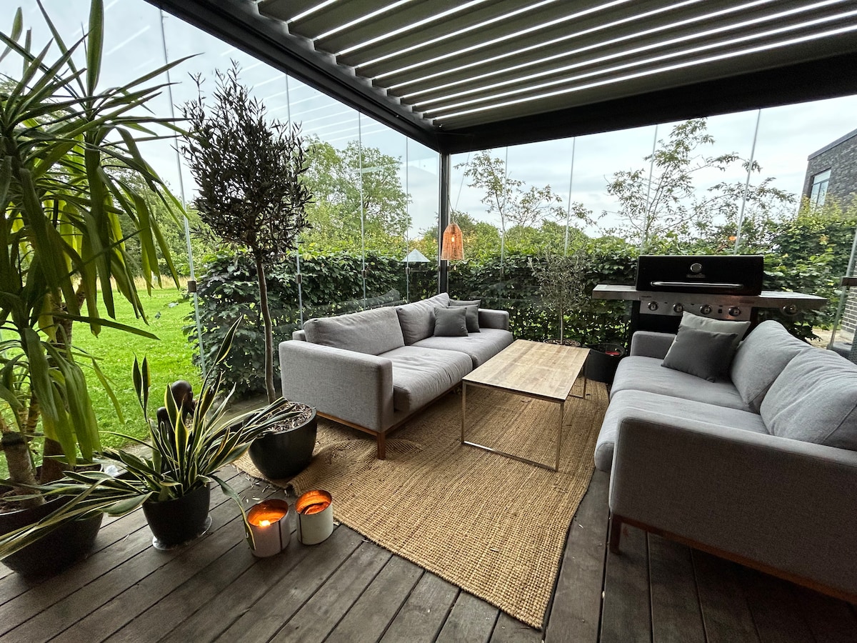 A covered outdoor seating area is furnished with two grey sofas and a modern coffee table. Potted plants and candles add to the ambiance. The background reveals a lush green garden, creating a serene retreat for relaxation and socializing.