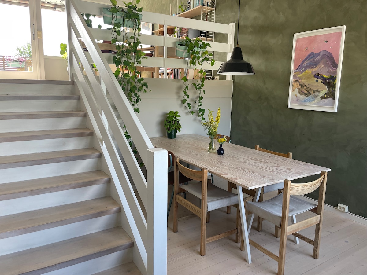 A wooden dining table with four chairs is positioned in a bright space, surrounded by greenery. A pendant light hangs above the table, and a colorful artwork is displayed on the wall. Natural light floods in from a nearby window, enhancing the atmosphere.