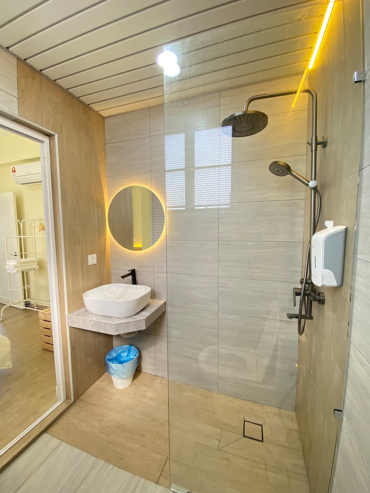 A modern bathroom is showcased, featuring a walk-in shower with a rainfall showerhead. A round mirror is mounted above a sleek sink, accompanied by a small countertop. Natural light enters through a window, and a waste bin is visible near the sink area.
