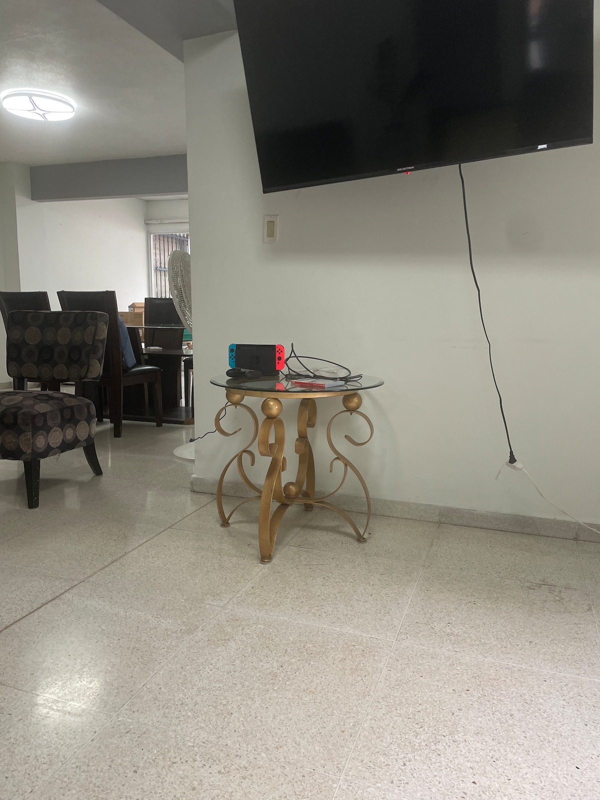 A stylish side table with a decorative base is positioned beside a television mounted on the wall. A gaming console is placed on the table, while a glimpse of dining chairs and a window is visible in the background, contributing to the open living space.