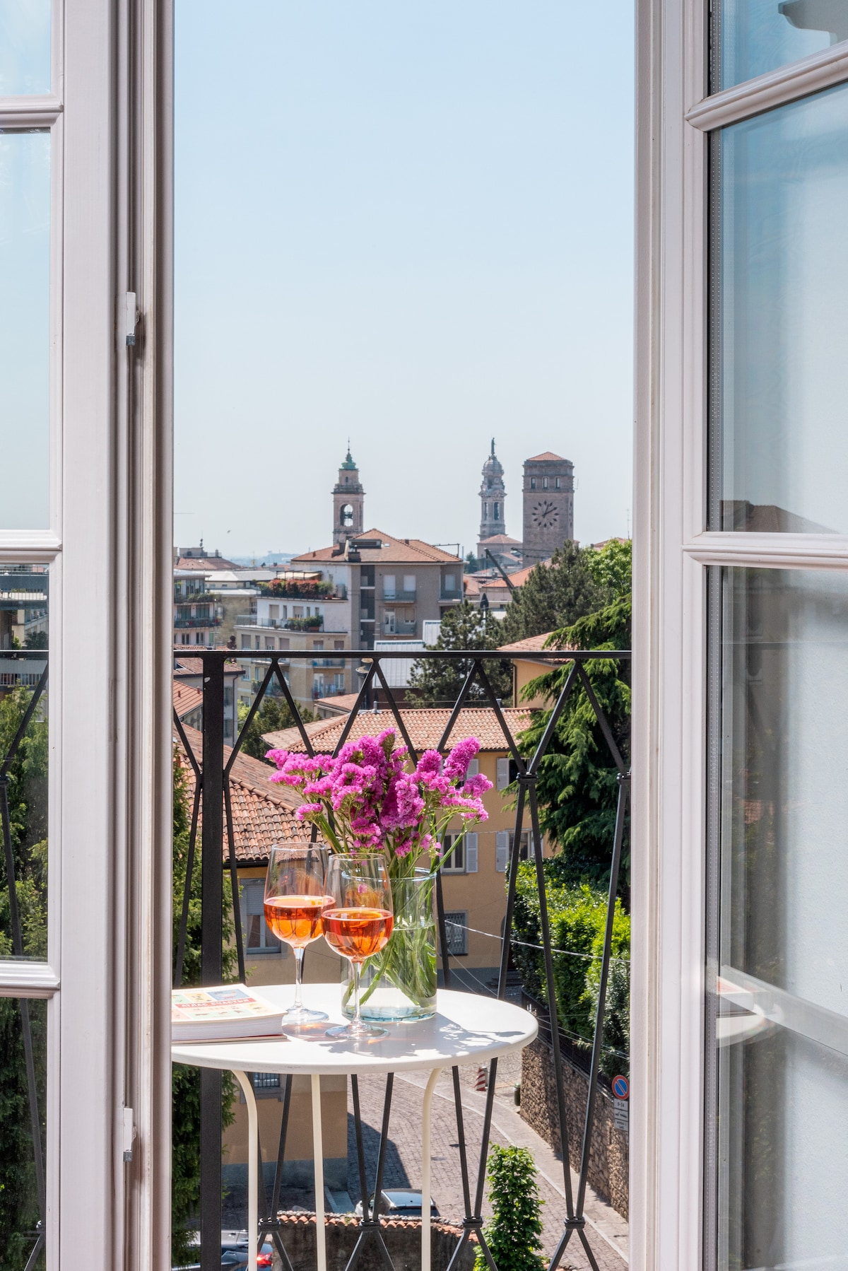 A beautiful view from an open window showcases a charming balcony with a small table set for two. Glasses of a refreshing drink sit next to a clear vase filled with vibrant flowers, with historical towers visible in the background.